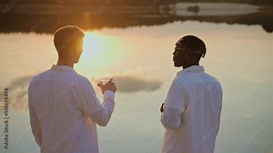 Handsome young men spend sunset at the beach, vaping and drinking. Friends chatting at the lake beach party. Black man vaping talking with a friend at the beach party. High quality 4k footage
