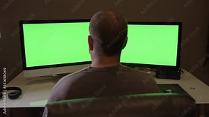 Behind man using dual screen computer at home office with green screen for keying.