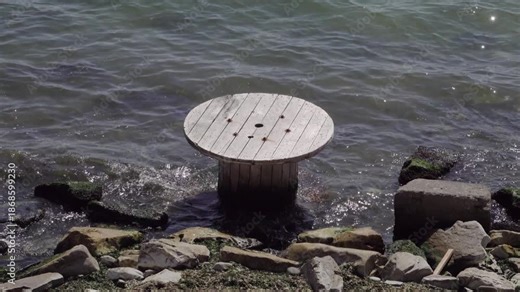 Large wooden cable spool lying in shallow water on the shore, representing environmental pollution, coastal waste and ecological issues affecting marine and shoreline areas