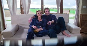 Husband and wife are watching a movie together, husband'd arm is over her wife's shoulder. He is changing the channel and she is giving him some chips. Wide-angle shot.
