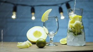 Close up cooking melon lemonade. Pouring soda water into a jug with pieces of melon, lime wedges, mint leaves and ice. Blurry lamps on blue background. Slide or move camera, dolly shot