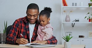 Afro american single father checking homework helping cute african school child daughter with studies teaches baby to write sitting at living room table together. Private education during lockdown
