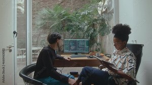 Young male and female programmers discussing computer codes and working together on IT project in home office with backyard garden view. Zoom-out shot