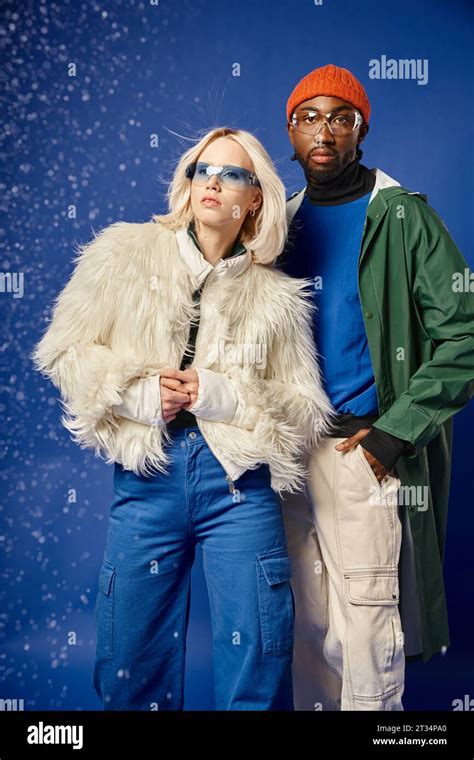 african american man and blonde woman posing in winter attire under falling snow on blue