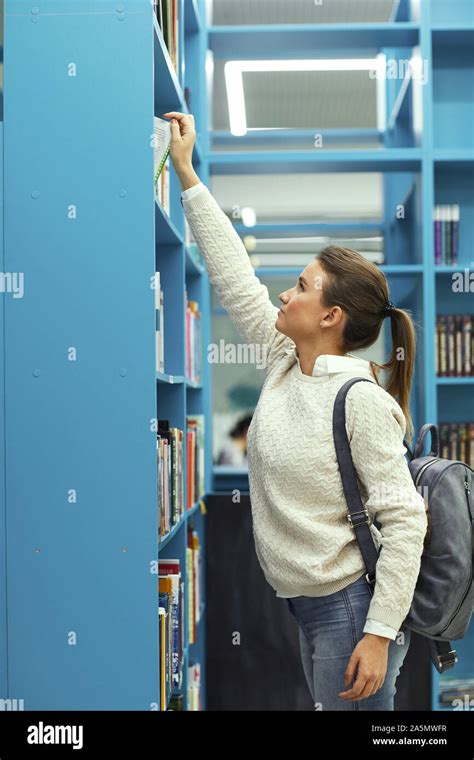 Side View Portrait Of Teenage Girl Reaching For Book At Top Shelf In