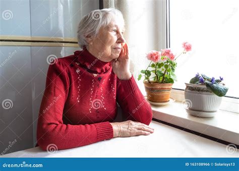 Old Lonely Woman Sitting Near The Window In His House Stock Photo