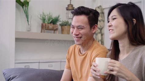 Asian Couple Watching Tv And Drinking Warm Cup Of Coffee In Living Room