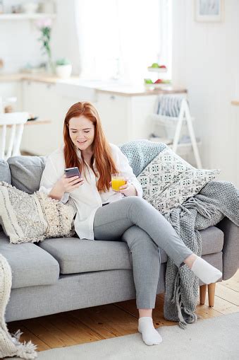 Smiling Young Woman With Red Hair Sitting On Sofa At Home And Reading