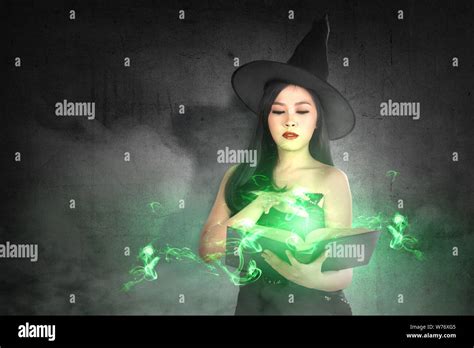 Asian Witch Woman In Hat Learns The Spell From The Magic Book With A Black Wall Background