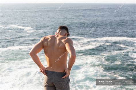Back View Of Muscular Male Athlete With Naked Torso Standing On Beach