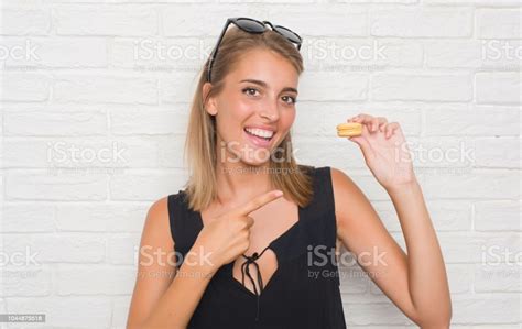 매우 행복 한 손과 손가락으로 가리키는 노란색 마카롱 먹고 흰 벽돌 벽에 아름 다운 젊은 여자 가리키기에 대한 스톡 사진 및 기타 이미지 Istock