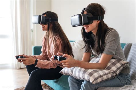 Two Women Sitting On A Couch Playing Video Games With Virtual Headsets Over Their Eyes