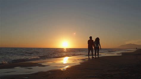 Young Couple Walking Along Beach In Sunset Holding Hands Free Video