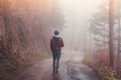 Man Walking Alone On A Road Through A Foggy Forest With Sunshine