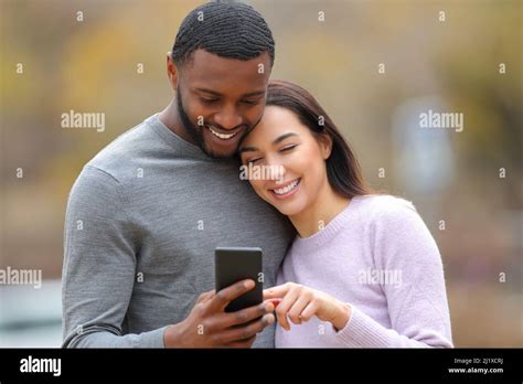 Front View Of A Happy Interracial Couple Checking Smart Phone Outdoors