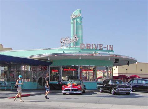 Two People Walking In Front Of A Drive In With Cars Parked On The Street