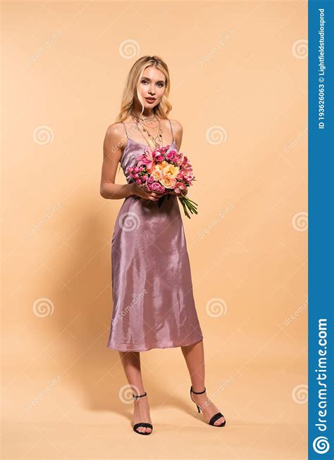 Blonde Woman In Violet Satin Dress Holding Bouquet Of Flowers On Beige