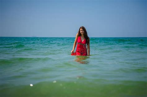Young Long Haired Brunette In Red Beach Dress Stands In The Turquoise Water Of The Ocean On A