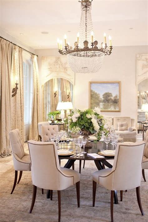 Sophisticated dining room with gray walls features a round wood antique dining table, white and heather gray french dining chairs with silver nailhead trim atop a white rug, white and pink abstract art over a gold leaf bar cart, window with blue and white roman shades and cream buffet cabinet. 33 Inviting And Cute Vintage Dining Rooms And Zones - DigsDigs