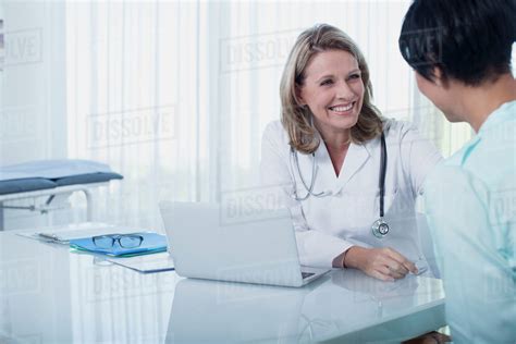 Smiling Female Doctor And Woman Sitting At Desk In Office Stock Photo