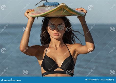 Portrait Of Gorgeous Dark Haired Girl In A Black Bra And Sunglasses Holds A Surfboard Over Her