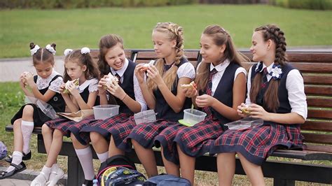 Girls College Student Wearing The Same School Uniform Eating Sandwiches Sitting On A Bench In