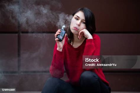 Vape 십대 겨울에 거리에서 현대 갈색 배경 맞은 편에 전자 담배를 흡연 빨간 스웨터에 젊은 예쁜 흰색 백인 소녀 나쁜 습관 전자담배에 대한 스톡 사진 및 기타 이미지