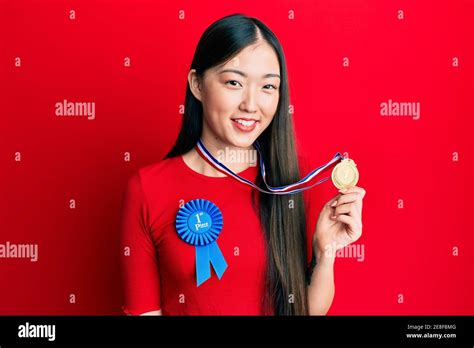 Young Chinese Woman Wearing First Place Badge Holding Medal Looking Positive And Happy Standing