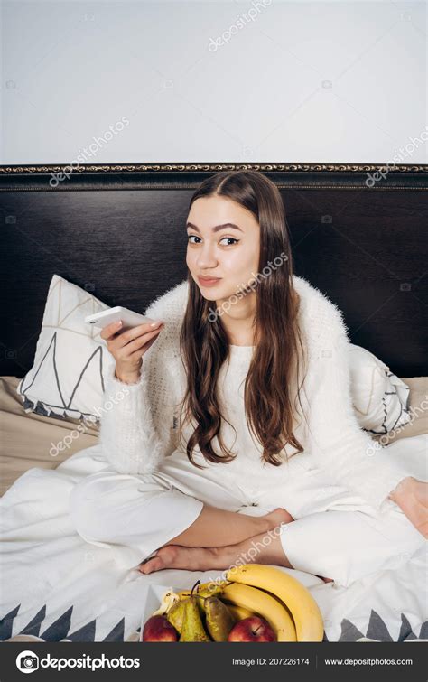 Funny Young Girl With Long Hair Sitting In Bed Early In The Morning Holding A Smartphone Stock