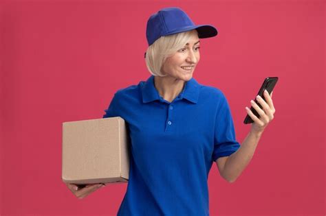 Free Photo Joyful Middle Aged Blonde Delivery Woman In Blue Uniform And Cap Holding Cardboard