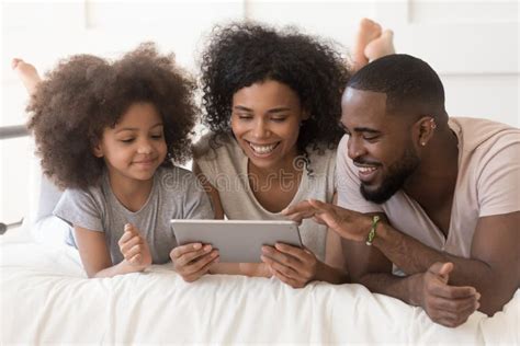 Happy African Parents With Daughter Using Tablet Lying On Bed Stock