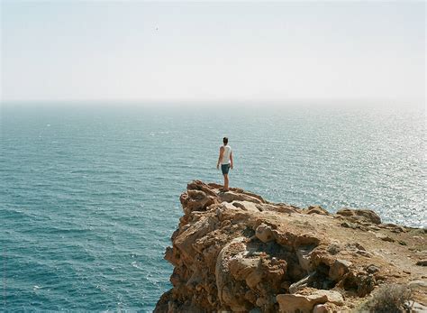 Man Standing On The Edge Of A Cliff Overlooking The Ocean By Stocksy