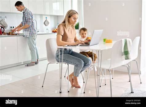 Busy Mother Working From Home Using Laptop Sitting At Table With Cute