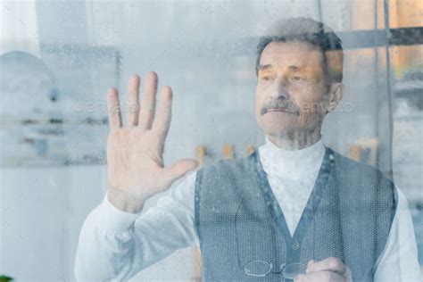 Sad Senior Man Waving Hand While Standing Near Window Stock Photo By