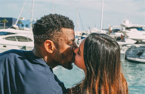 Close Up Of Interracial Couple Kissing At The Port With Boats Background In Summer During