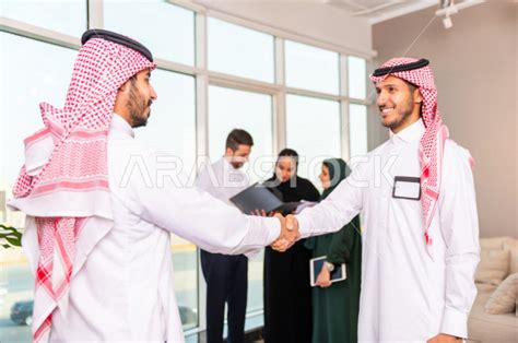 رجل اعمال سعودي خليجي يصافح شريكه في العمل ، و بالخلفية مجموعة من الرياديون السعوديون يناقشون