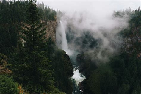 Waterfall In Mountainous Terrain With Evergreen Forest Against Cloudy