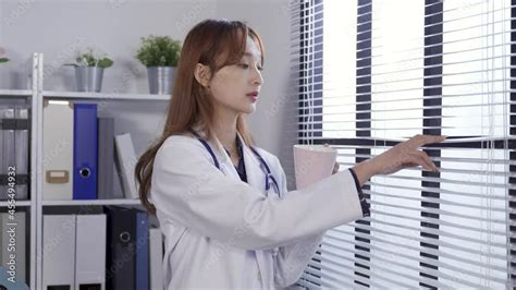 asian beautiful physician is parting the louver blinds with her fingers and watching outside