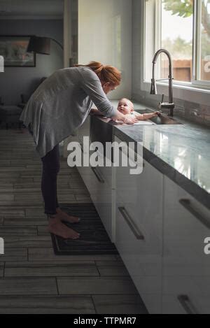 Side View Of Mother Bathing Cute Son In Kitchen Sink At Home Stock