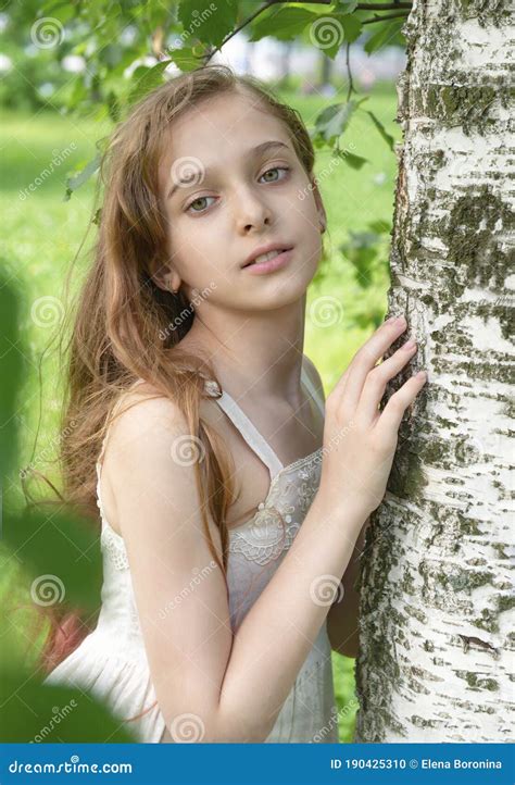 white beautiful girl 11 years old with long hair in a white dress near a birch tree on a green
