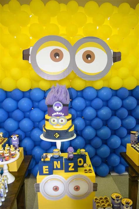 A Table Topped With A Cake Next To A Minion Balloon Wall Covered In