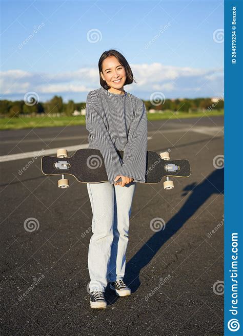 Vertical Shot Of Skater Girl Posing With Longboard Cruising On Empty Road In Suburbs Smiling
