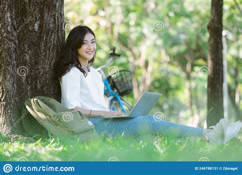 Young Asian College Girl Relax Sit On Grass With Ground Lean Big Old Tree At Park Browsing