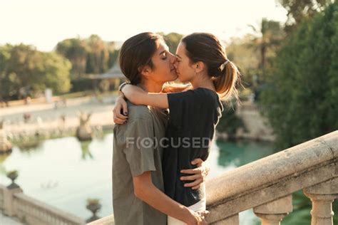 Portrait Of Boyfriend Embracing Girlfriend And Kissing In Nose On Stairs In Park Over Lake On