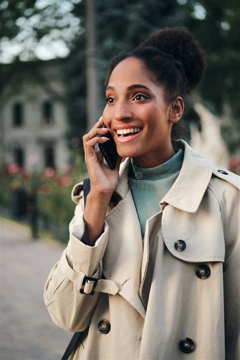 Pretty Emotional African American Girl In Trench Coat Happily Talking On Cellphone Outdoor Stock