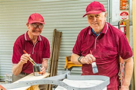 Image Of Two Elderly Handymen Smiling While Painting Ironwork Together In A Men S Shed