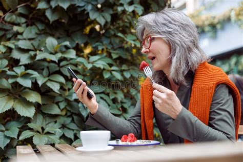 Mature Woman With Glasses Eats Dessert And Looks At Smartphone At Table