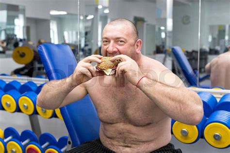 Big Fat Hungry Man Chewing A Hamburger With Meat And Cheese In The Gym