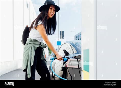 Young Woman Wearing Hat Charging Electric Car At Vehicle Station Stock