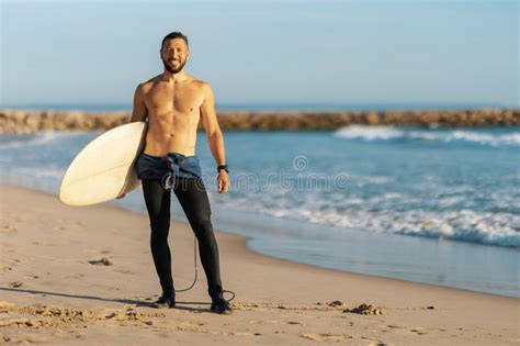 A Man Surfer With Naked Torso Standing On The Seashore Holding A